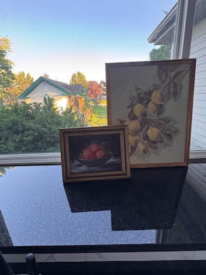 Uma mesa com duas pinturas emolduradas e vista para uma casa e árvores pela janela.