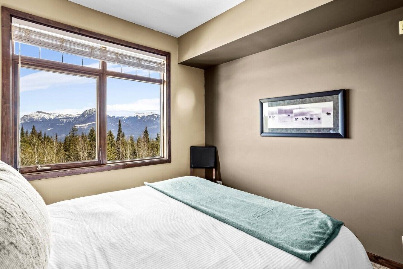 A bedroom with a large window offering a mountain view, a bed with a white and blue blanket, and a framed picture on the wall.