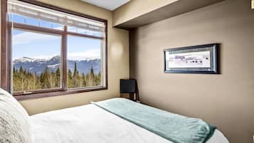 A bedroom with a large window offering a mountain view, a bed with a white and blue blanket, and a framed picture on the wall.