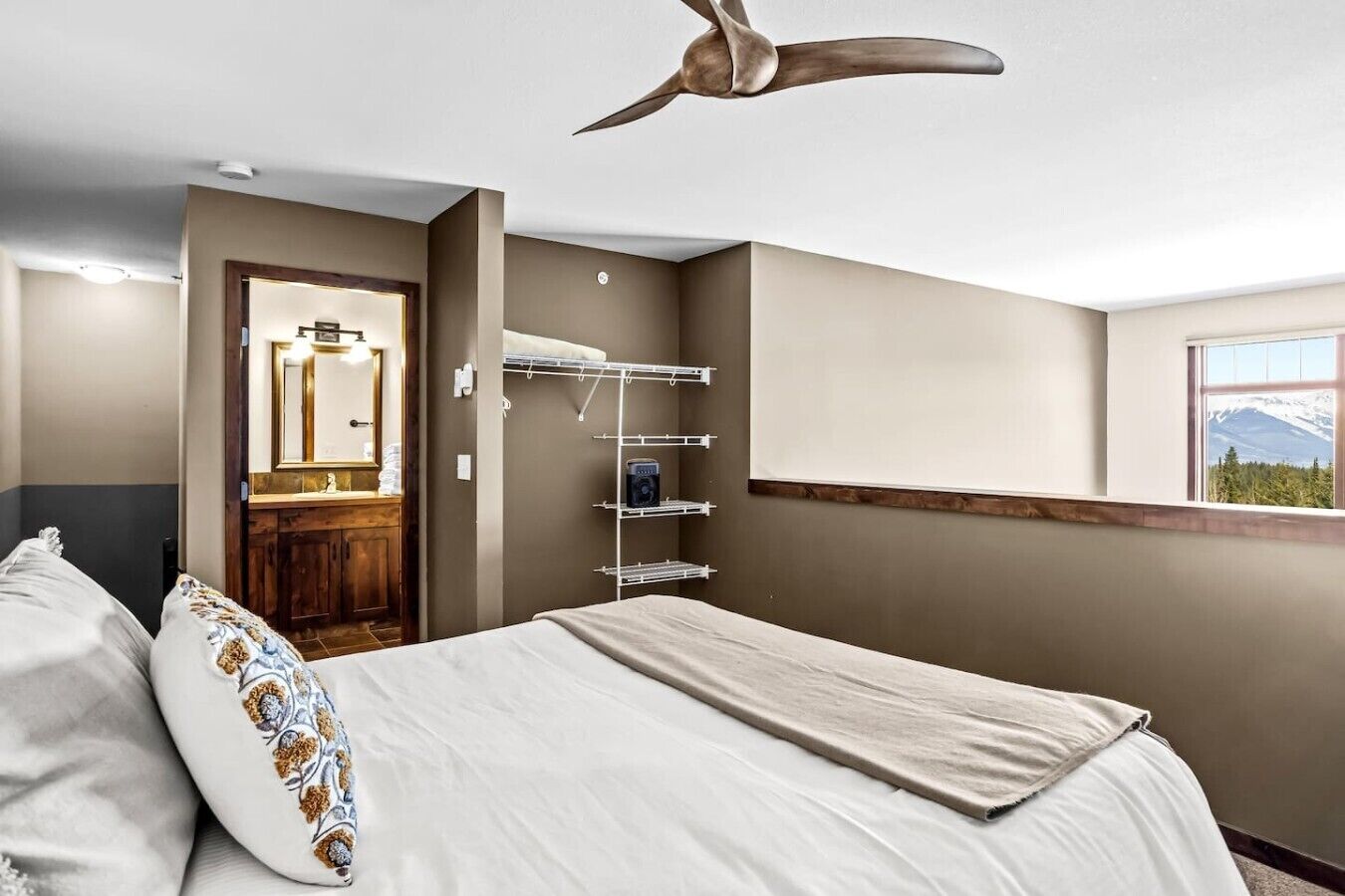 Une chambre à coucher comprenant un lit, un ventilateur de plafond, une étagère intégrée, une fenêtre donnant sur la montagne, ainsi qu’une salle de bain avec un miroir et un lavabo.
