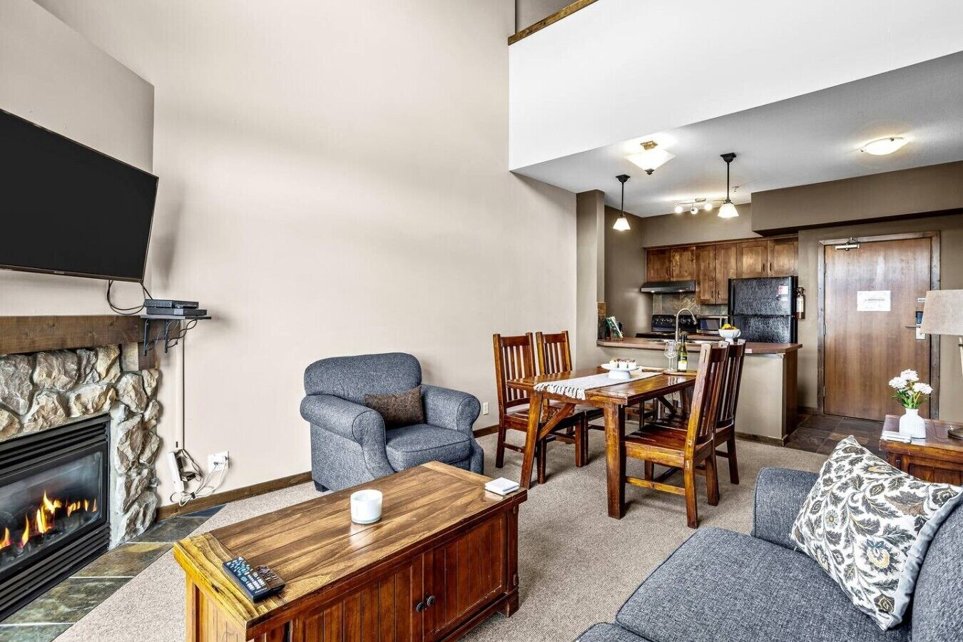 Un salon avec une cheminée, un téléviseur fixé au mur, un coin repas avec des meubles en bois et, au fond, une cuisine.