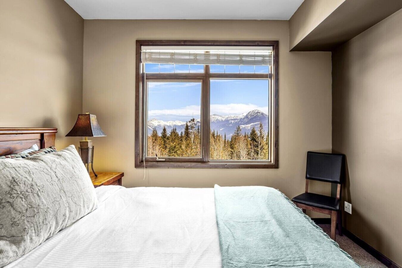A bedroom with a bed, a chair, a nightstand, a lamp, and a window with a mountain view.