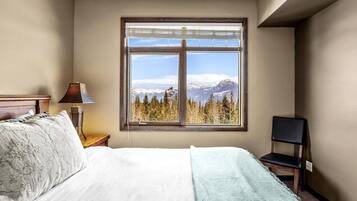 A bedroom with a bed, a chair, a nightstand, a lamp, and a window with a mountain view.