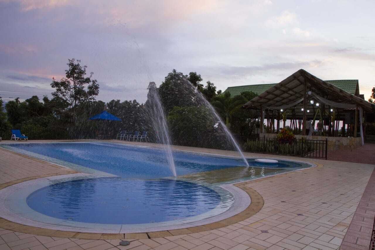 Una piscina con una fuente de agua, rodeada por un solarium con baldosas y un pabellón cubierto.