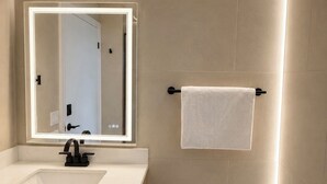 Ein modernes Badezimmer mit einem weißen Waschbecken, einem Spiegel mit integrierter Beleuchtung, einem Handtuchhalter, einer Toilette und einem Mülleimer.