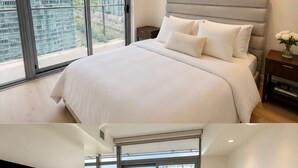 Ein modernes Schlafzimmer mit einem großen Bett, einem Nachttisch, einem Fernseher und Blick auf eine Skyline durch große Fenster.