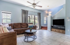 A living room with a sectional sofa, a round coffee table, a ceiling fan, a flat-screen TV, and large windows. - Disney Area Family Oasis (Kissimmee)