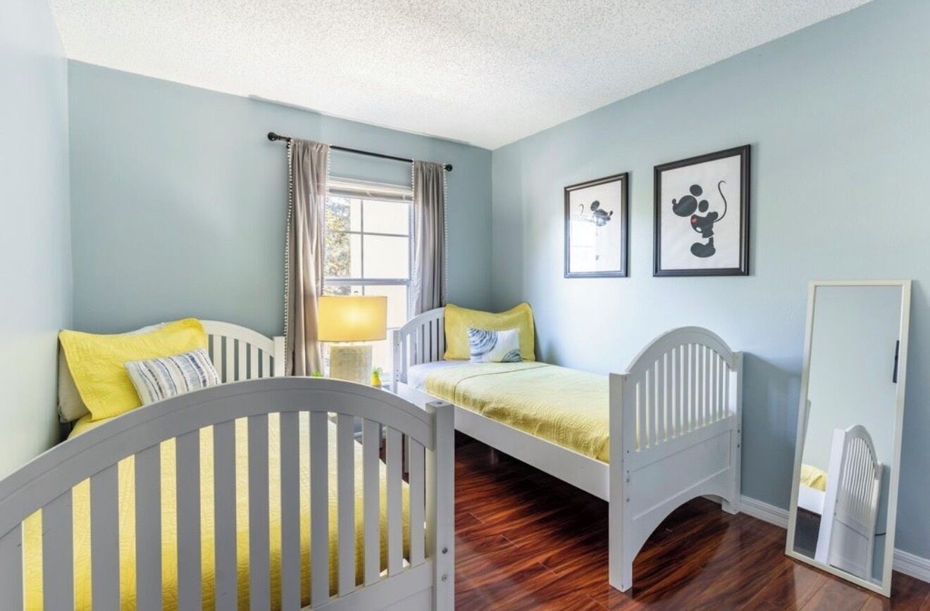 A room with two beds, a crib, a window with curtains, and framed pictures on the wall.