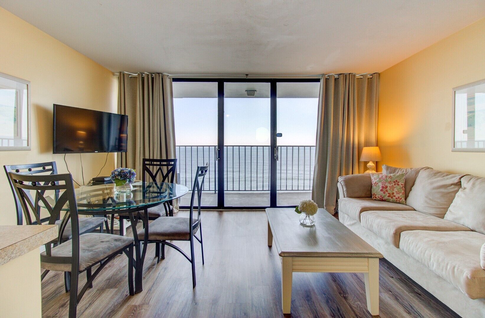 A hotel room with a balcony, a dining area with a glass table, a sofa, and a TV.