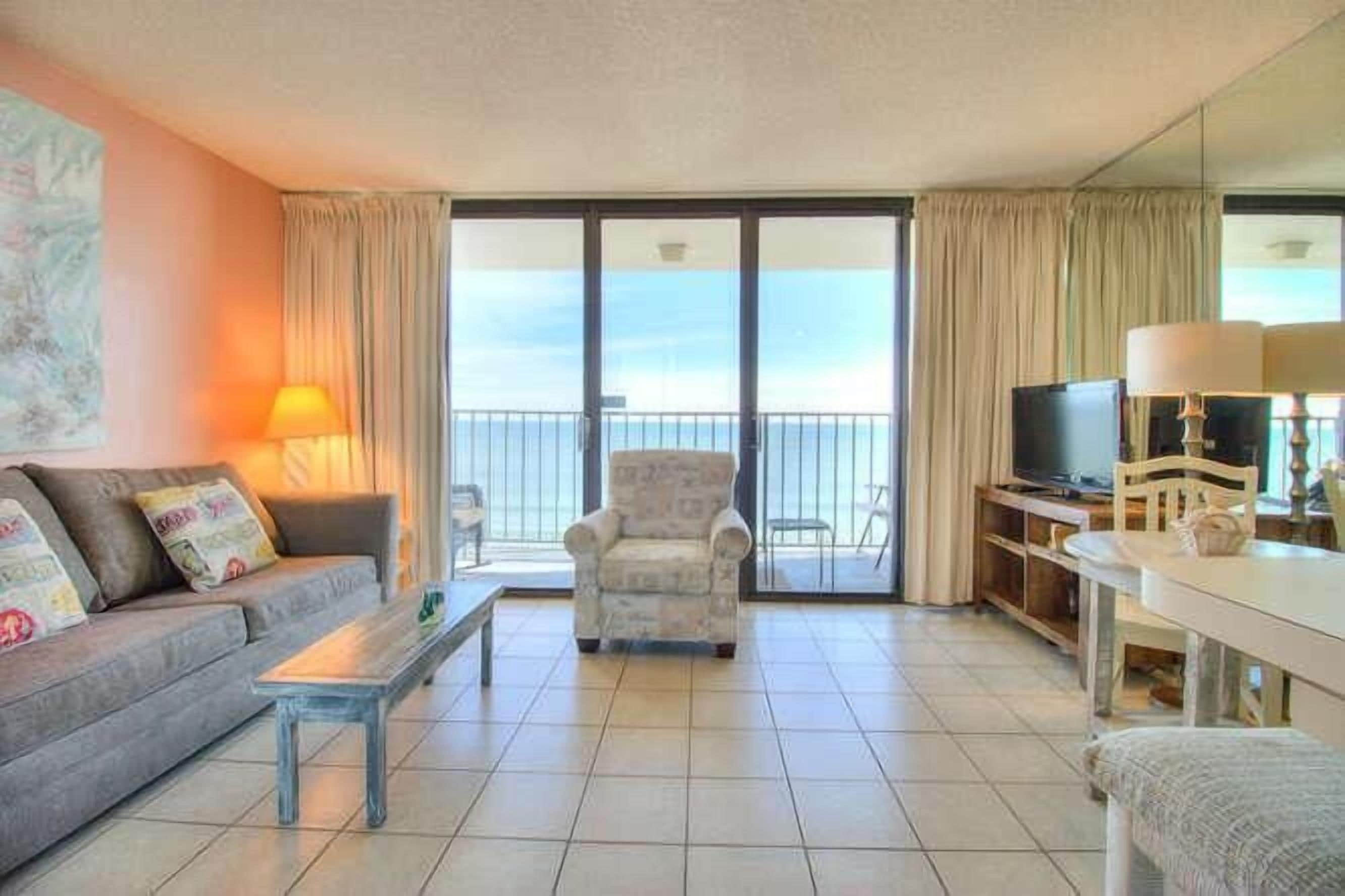 A living room with a sofa, armchair, TV, and a balcony with curtains.