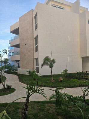 A modern building with a curved pathway, a small palm tree, and various plants. - 2BR 2BA Family Friendly Beachfront Oasis in Brand New Development! (Cabarete)