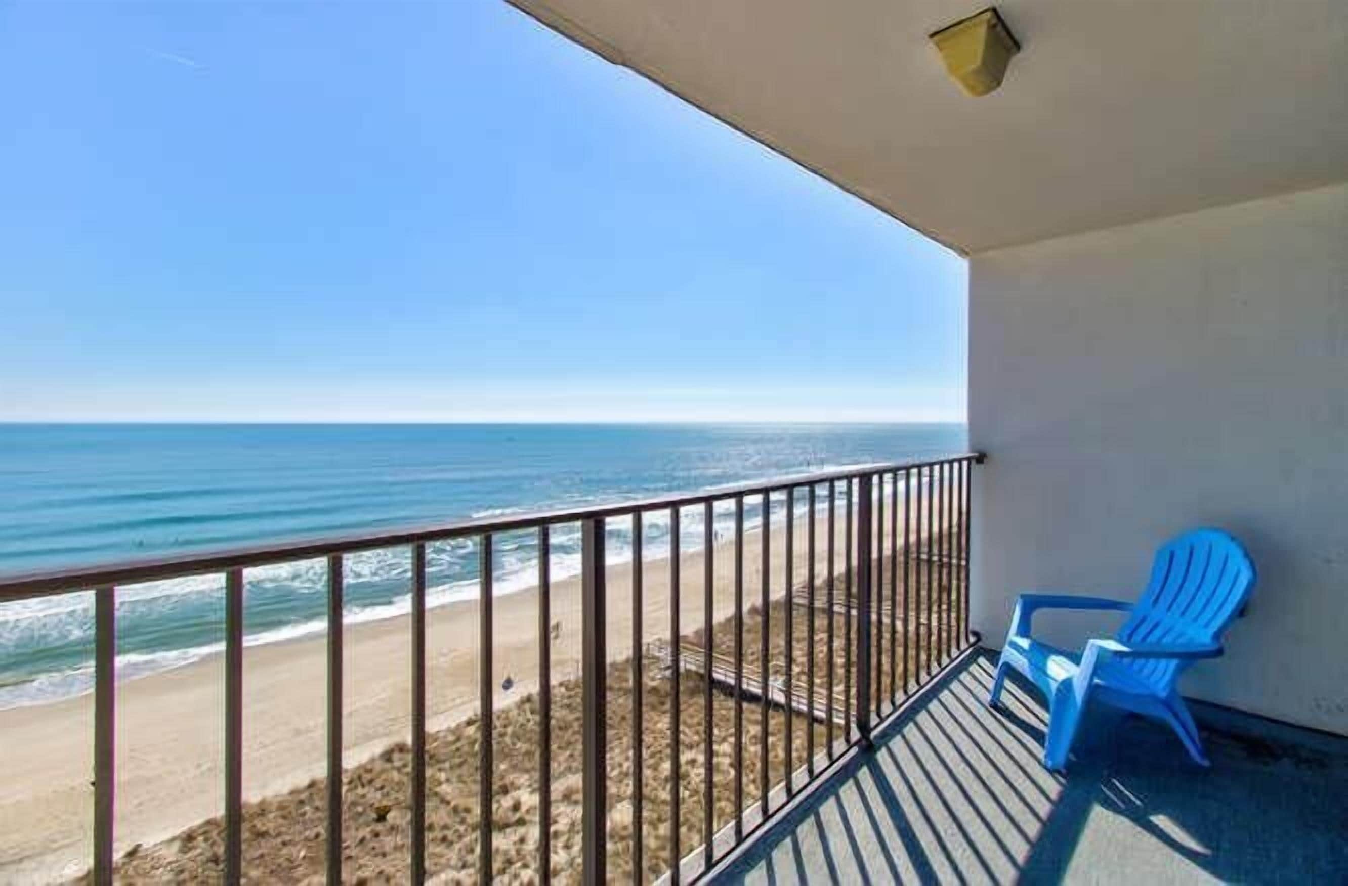A balcony with a view of the beach and ocean, a blue chair, and a railing.