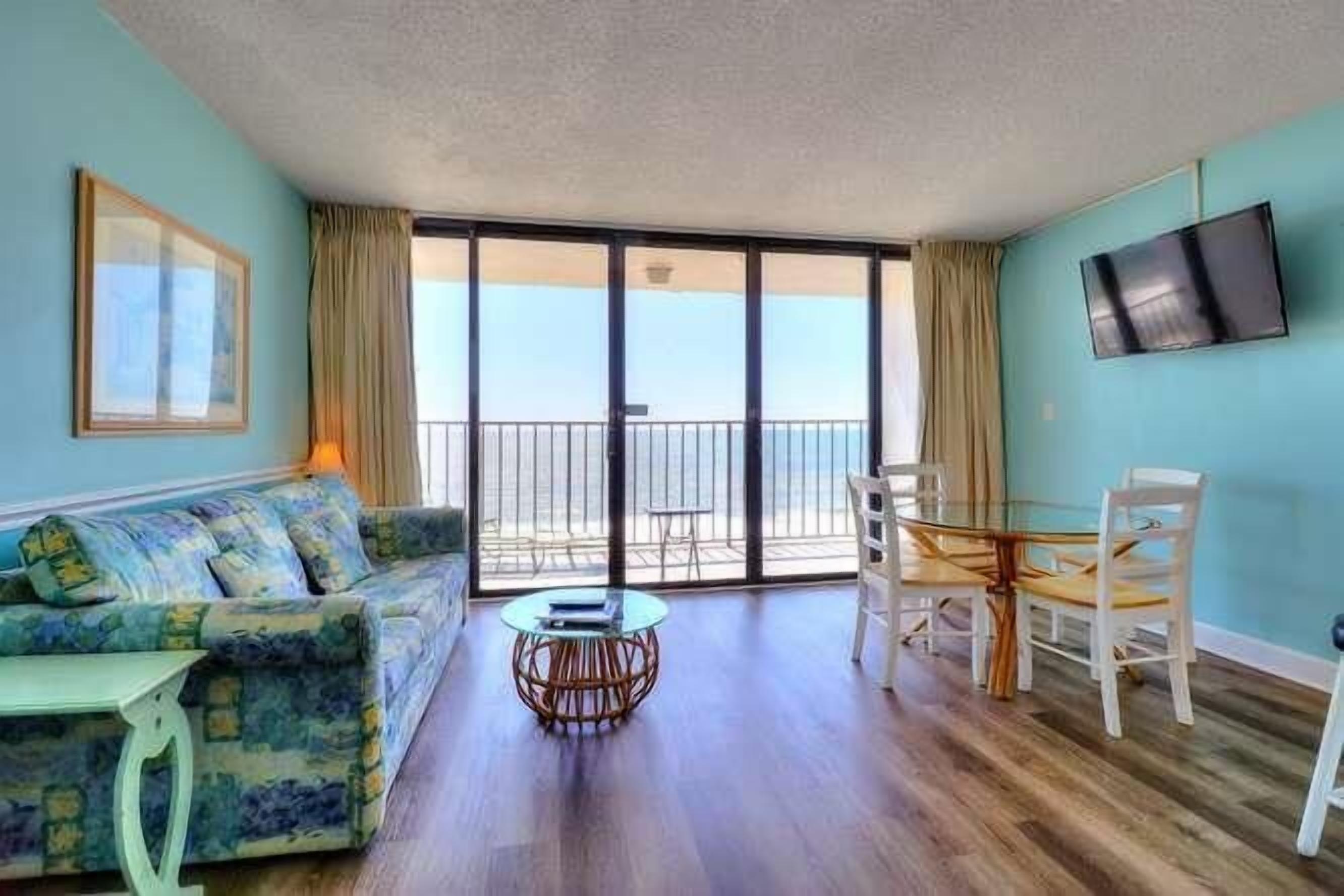 A room with a sofa, a dining table, chairs, a TV, and a balcony with a view.