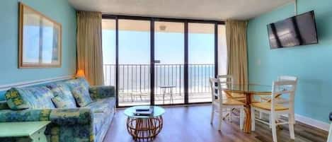 A room with a sofa, a dining table, chairs, a TV, and a balcony with a view.