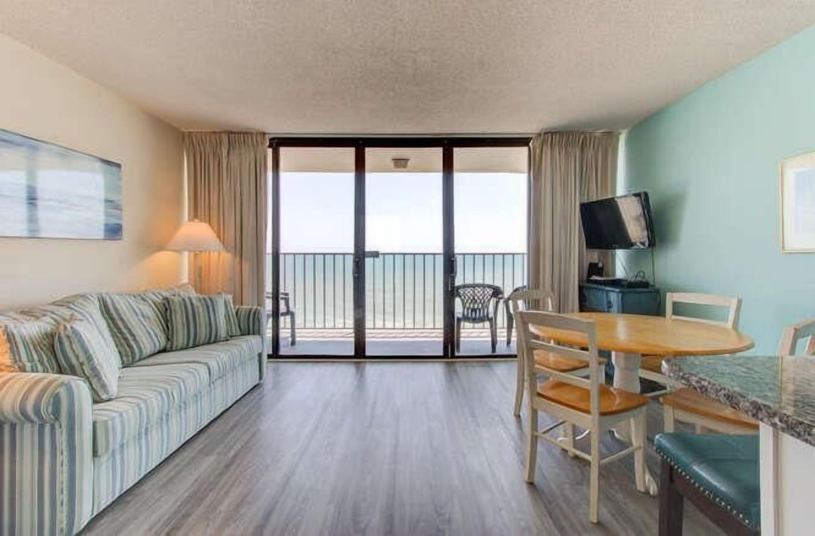 A hotel room with a sofa, dining table, chairs, a TV, and a balcony with a view.