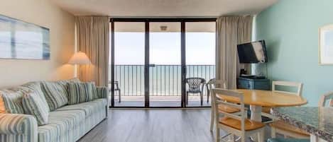 A hotel room with a sofa, dining table, chairs, a TV, and a balcony with a view.