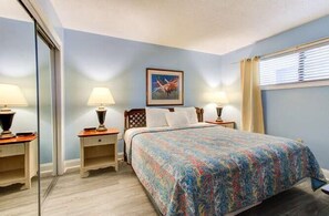 A bedroom with a bed, two bedside tables with lamps, a framed picture on the wall, and a window with blinds. - Oceanfront 1BR condo in Carolina Beach 0909 (Carolina Beach)