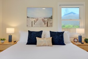 A neatly made bed with two dark blue pillows and a beige pillow with 'Seas the Day' text, flanked by two bedside lamps and a small plant on a wooden nightstand. - NEW! Sea Breeze Cottage - Remodeled, A/C, Indoor/Outdoor Retreat (Santa Barbara)