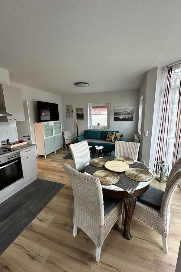 A modern kitchen with white cabinets, a dining area with wicker chairs and a round table, and a living area with a teal sofa. - FEWO „Am Stausee“ by Interhome (Friesoythe)