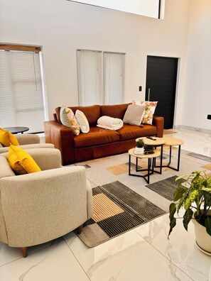 Una sala de estar moderna con un sofá de cuero marrón, un sillón beige, una mesita redonda y una planta en maceta.