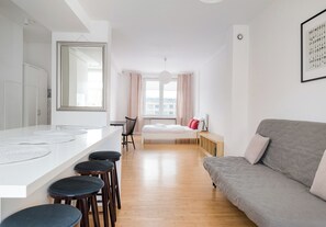 A modern, well-lit room with a large mirror, a sofa, a bed, a dining area with stools, and a view of buildings through the window. - Żelazna Urban Comfort Apartment (Warszawa)