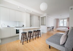 A modern kitchen with white cabinets, a central island with stools, a dining area with a table and chairs, and a gray sofa in a living area. - Żelazna Urban Comfort Apartment (Warszawa)