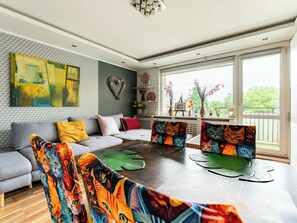 A modern living room with a grey sofa, colorful cat-themed cushions, a wooden coffee table, and large windows with a view of greenery.