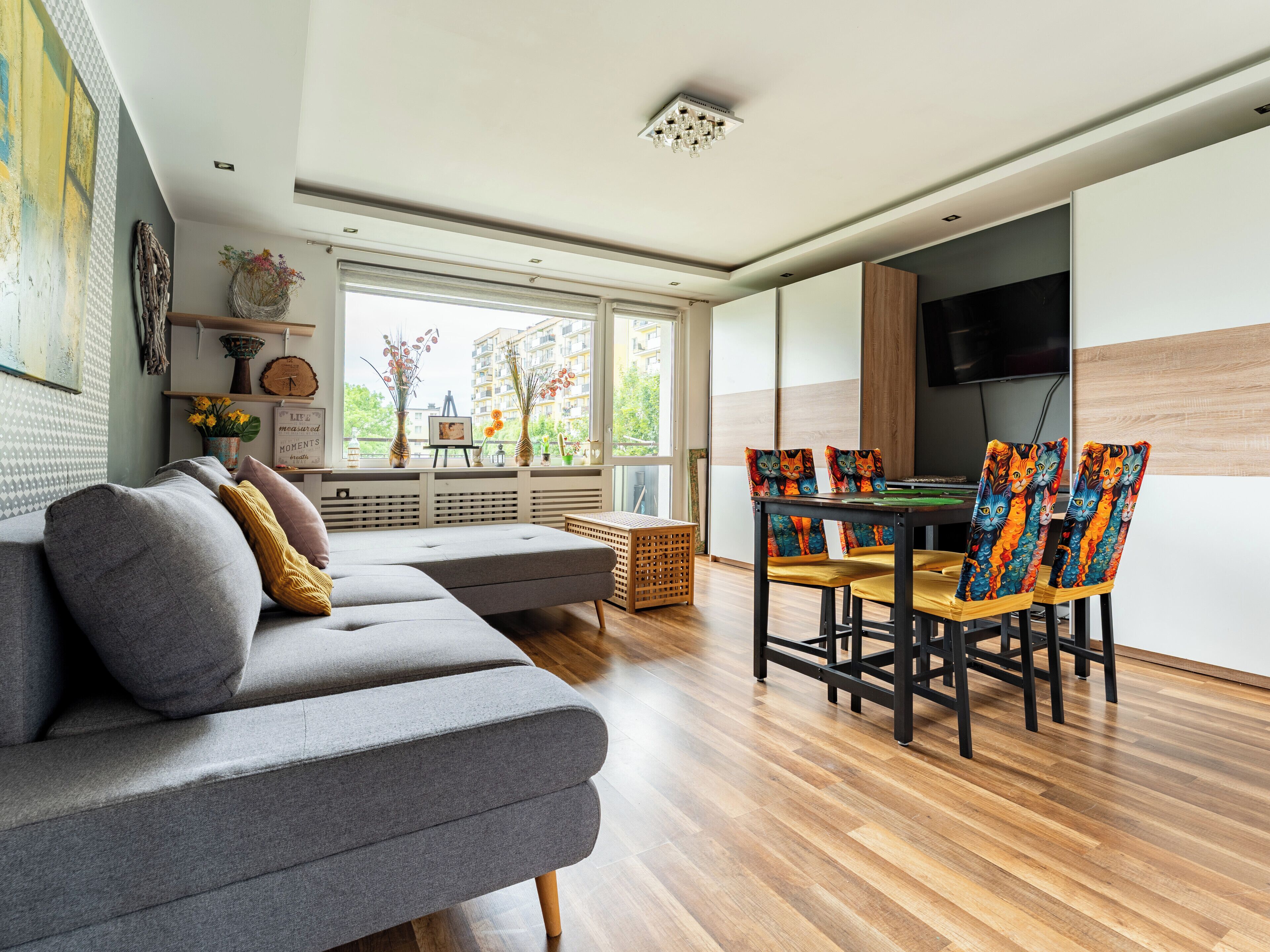 A modern living room with a grey sofa, wooden dining table, and colorful cat-themed chairs.