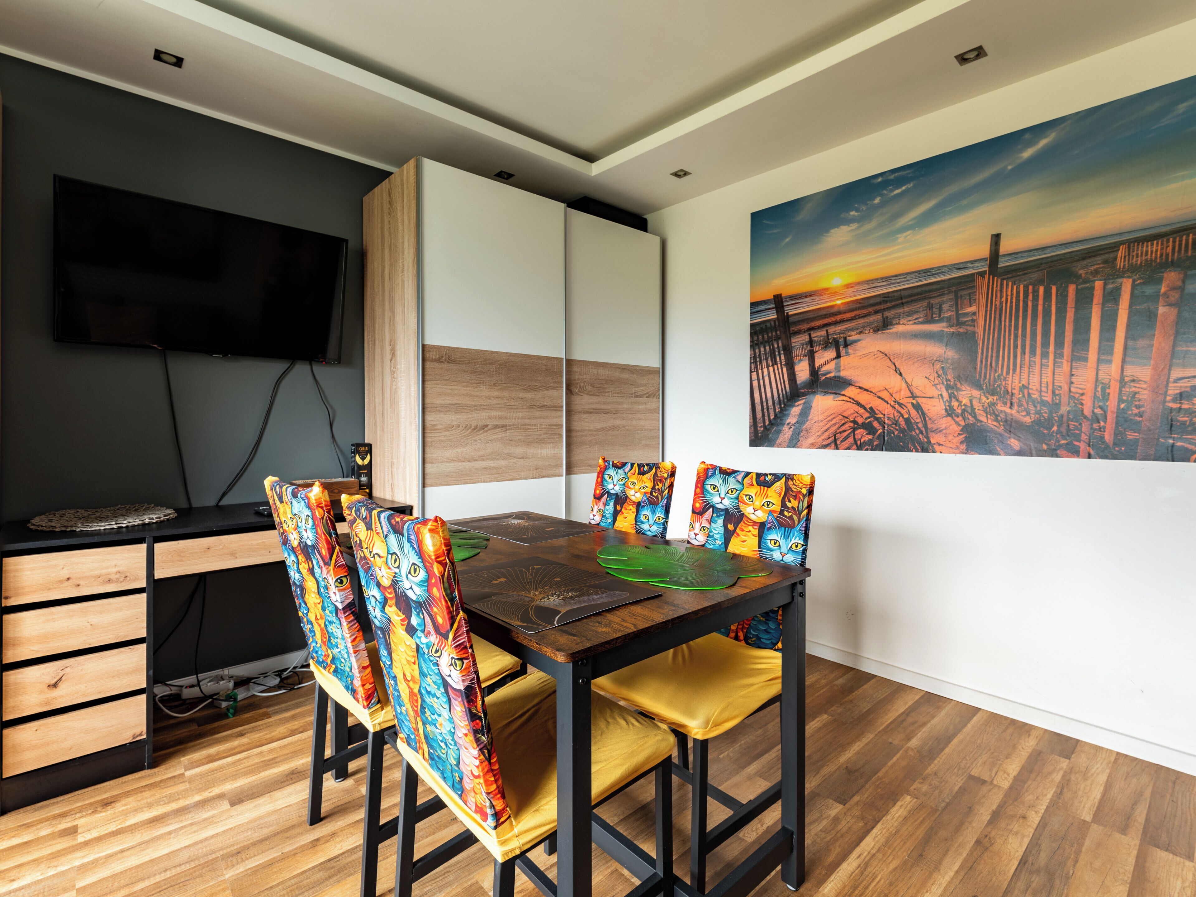 Un coin repas avec une table en bois, des chaises dotées de coussins colorés, un téléviseur fixé au mur et un tableau représentant un coucher de soleil.