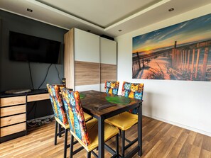 A dining area with a wooden table, chairs with colorful cushions, a television mounted on the wall, and a sunset painting on the wall.