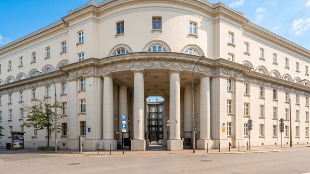 Un grand bâtiment de style classique, doté d’une entrée centrale flanquée de colonnes, sous un ciel dégagé, et donnant sur une rue pavée.