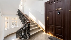 Un escalier avec une rampe en métal noir menant à un palier, une porte en bois sombre avec une plaque numérotée, et une fenêtre à carreaux multiples.