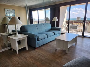 Ein Zimmer mit einem blauen Sofa, zwei weißen Nachttischen mit Lampen, einem weißen Couchtisch und Meerblick durch große Fenster.