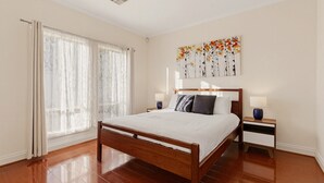 A bedroom with a wooden bed, a nightstand with a lamp, a window with curtains, and a wall art piece. - Family-friendly 4-bed Home With Parking (Adelaide)