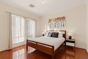 A bedroom with a wooden bed, a nightstand with a lamp, a window with curtains, and a wall art piece. - Family-friendly 4-bed Home With Parking (Adelaide)