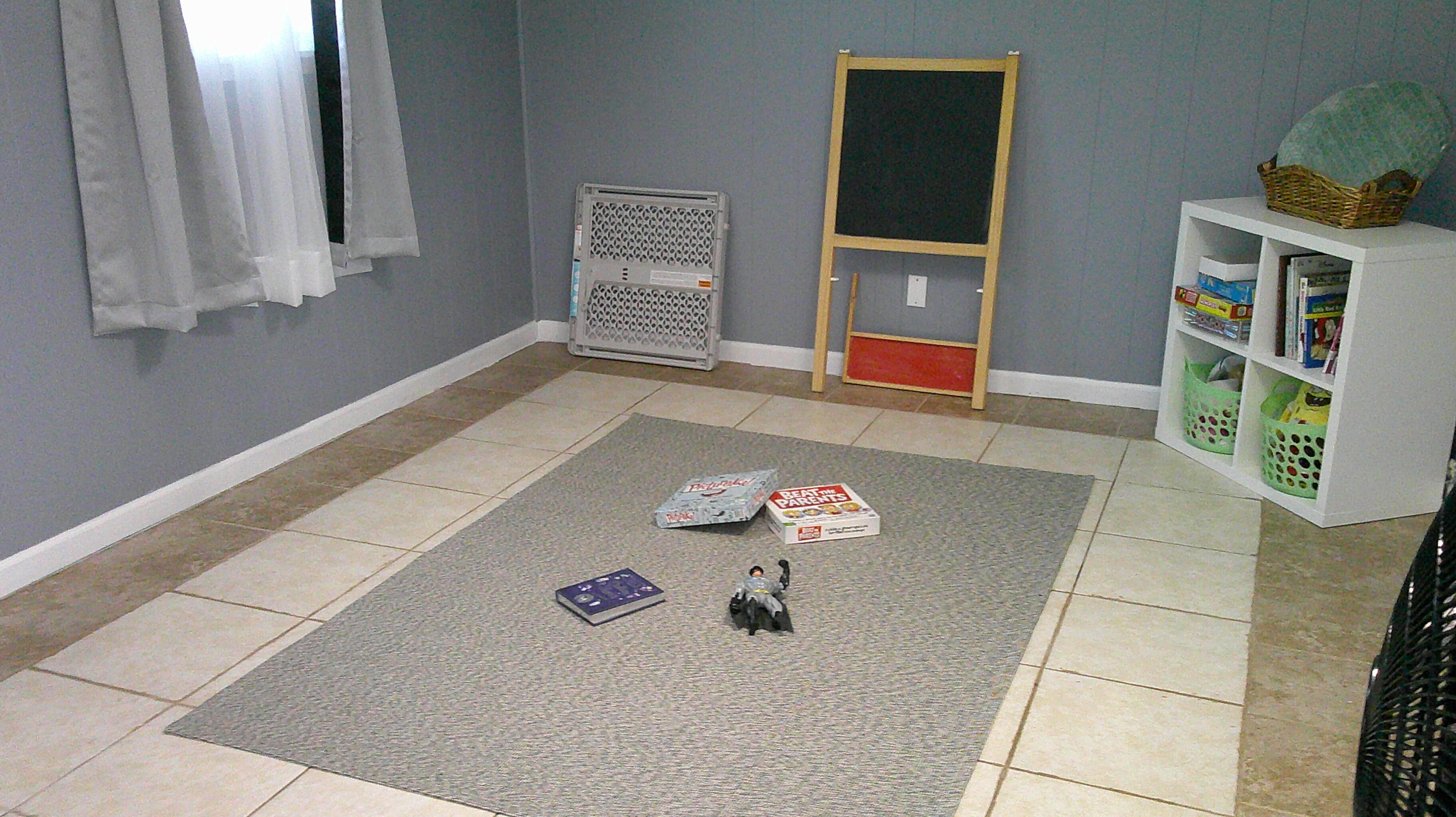 A room with a grey carpet, a white radiator, a bookshelf with baskets, and a small play area with toys.
