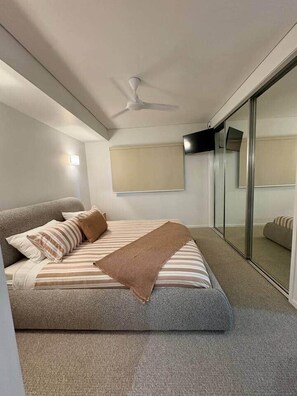 Ein Schlafzimmer mit Bett, Deckenventilator, Schiebeglasfenster und einem an der Wand montierten Fernseher.