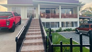 Un edificio de dos pisos con un balcón, una escalera que lleva a la entrada y un camino de acceso con vehículos estacionados.
