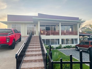 A two-story building with a balcony, a staircase leading to the entrance, and a driveway with parked vehicles. - Hearthstone Suite & Cafe (Laguindingan)