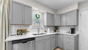A kitchen with grey cabinets, a white countertop, a dishwasher, a window with a wreath, and a coffee maker. - Walk to Martin City + Sleeps 6 + Yard + Fire Pit (Kansas City)