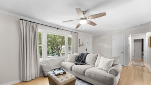 A living room with a sofa, a wooden ottoman, a ceiling fan, and a view of the outdoors through large windows. - Walk to Martin City + Sleeps 6 + Yard + Fire Pit (Kansas City)