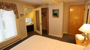 Ein Hotelzimmer mit Bett, Nachttischlampe, Schrank, Bad mit Waschbecken und Fenster mit Jalousien.