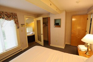Ein Hotelzimmer mit Bett, Nachttischlampe, Schrank, Bad mit Waschbecken und Fenster mit Jalousien.