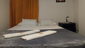 A hotel room with a bed, two white pillows, a dark gray bedspread, two towels, a wooden curtain, a nightstand with a lamp, and a painting on the wall.