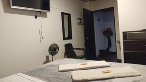 A bedroom with a bed, a TV mounted on the wall, a fan, and a door leading to another room.