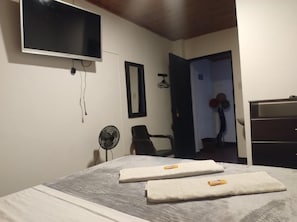 Un dormitorio con cama, televisor montado en la pared, ventilador y puerta que da a otra habitación.