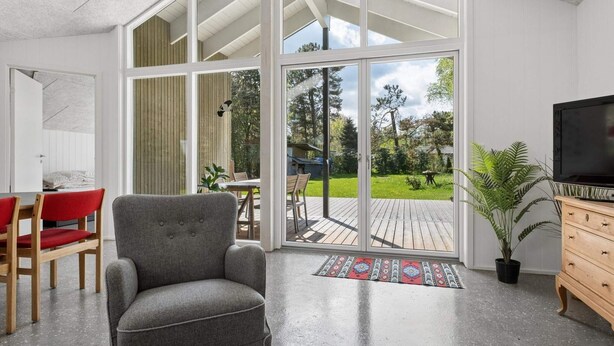 A modern living room with a grey armchair, wooden dining table, and chairs, a television, and a sliding glass door leading to a deck. - 4 Star Holiday Home in Storvorde (Storvorde)