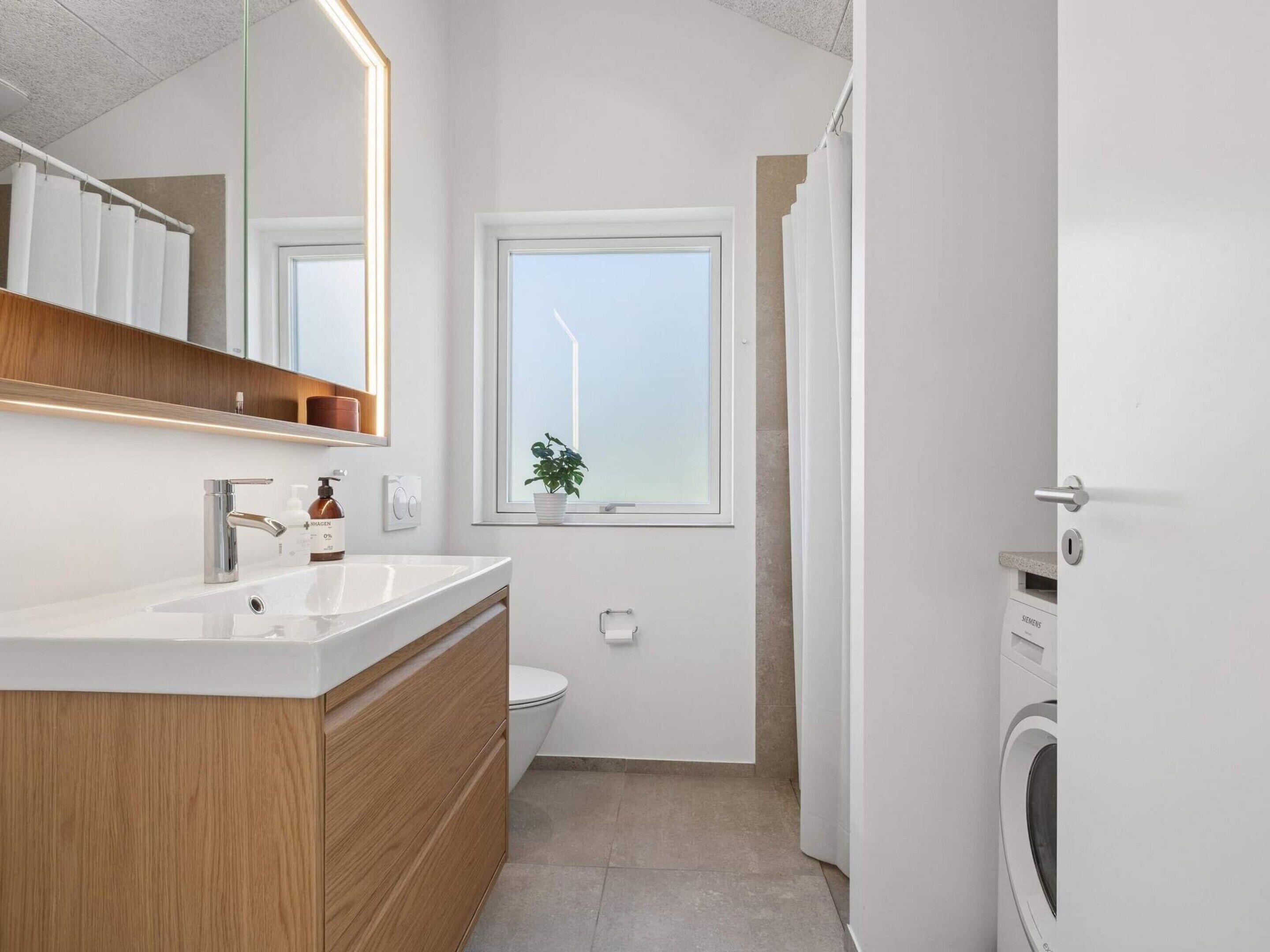 A modern bathroom with a wooden vanity, a white sink, a mirror, a window with a view, and a washing machine.