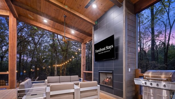 A wooden deck with a sofa, a TV mounted on a wall, and a grill. - NEW! Heated Pool, Hot Tub, Outdoor Lounge, Games (Broken Bow)