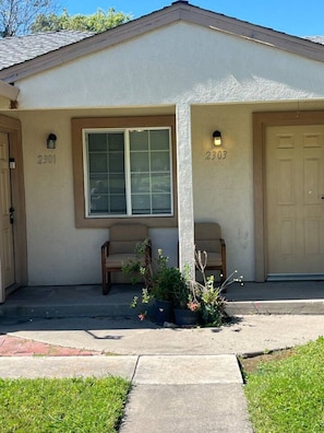 A single-story house with two garage doors, a window with blinds, and a small porch with a bench and potted plants.